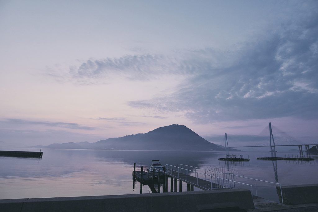 A tranquil evening view of the Seto Inland Sea, with Tatara Bridge and distant islands silhouetted against a pastel-colored sky.
